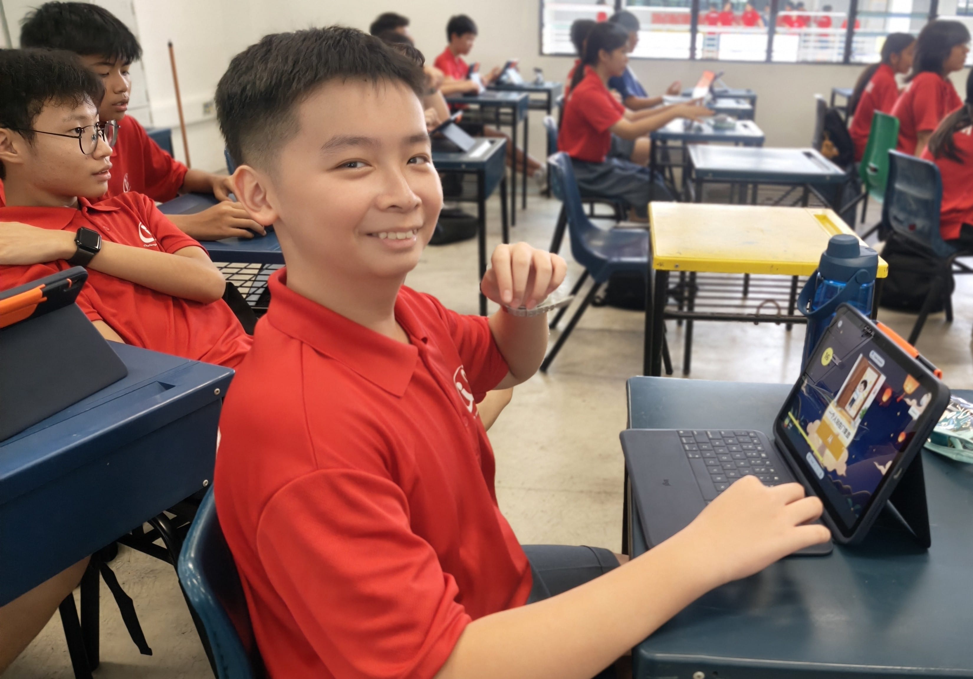 A secondary school boy in a red polo shirt sits at his desk, smiling at the camera while using a tablet displaying what appears to be a Mid-Autumn Festival digital learning activity. Other students working on tablets are visible in the background.